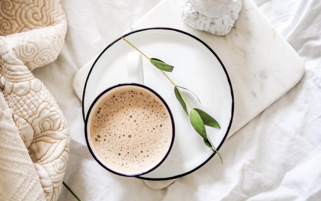 A cup of frothy coffee on a white plate with a green sprig on white blanket